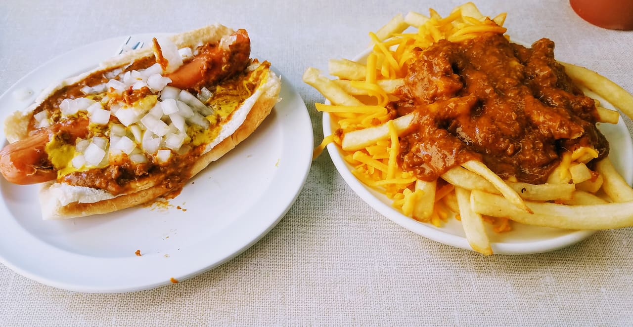 A coney dog under beef chili, yellow mustard and chopped white onion, beside a plate of chili cheese fries.