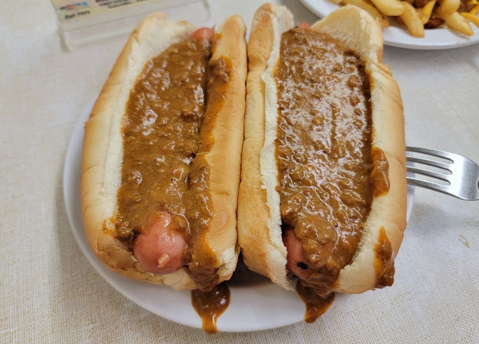 Two coneys on a white oval plate, buried under beef chili, with a plate of chili cheese fries behind.
